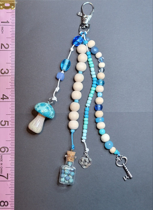 A handmade beaded keychain charm set displayed on a dark gray background next to a pink measuring ruler for scale. The keychain features cream-colored wooden beads, light blue and dark blue beads, a blue and white marbled mushroom charm, a small glass vial with blue beads, and a silver key.