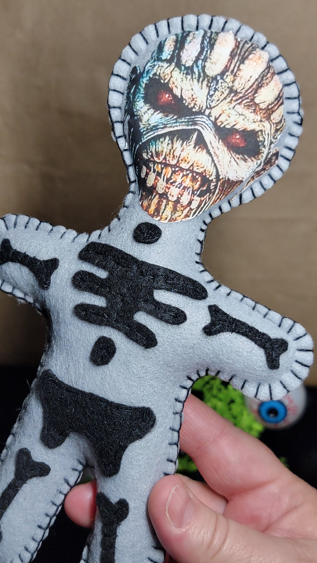 A handmade gray felt voodoo doll with black felt skeleton patterns and a printed skull face, held by a hand against a brown background.