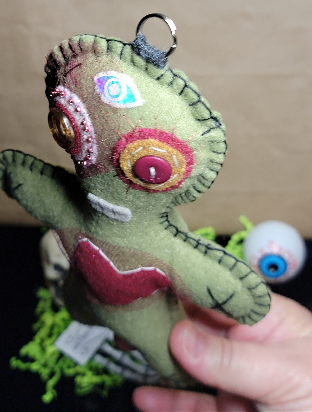 A handmade green felt soul doll keychain with colorful embroidered eyes and a red stitched heart, held by a hand against a neutral background.