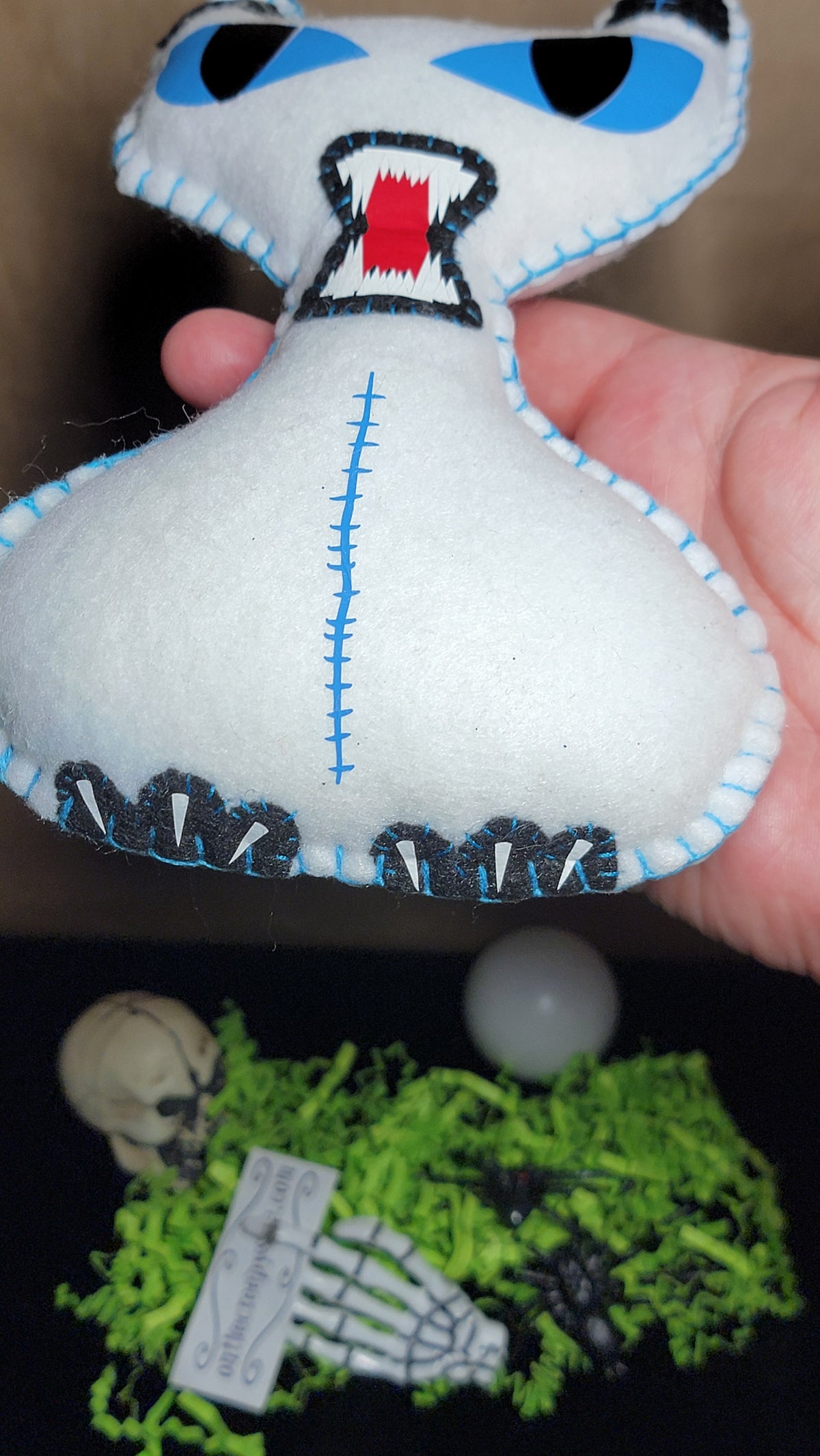 A handmade white felt monster doll with blue eyes, a stitched mouth, and black and white teeth, held by a hand over a themed background with green moss, a small skull, and skeleton decorations.