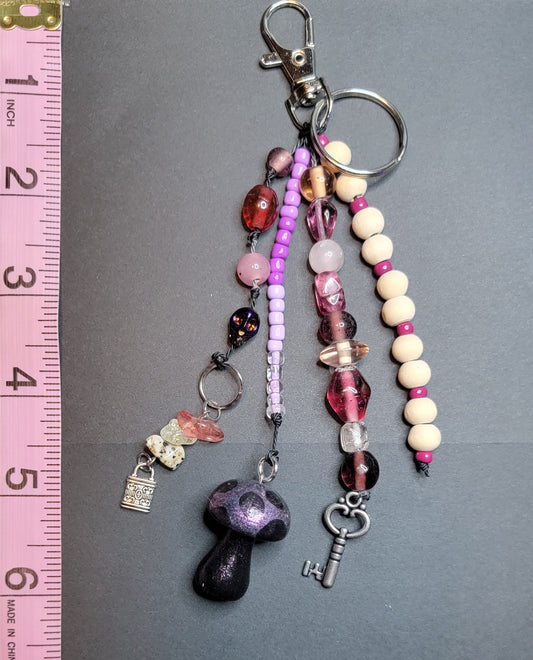 A beaded keychain with charms, including a purple mushroom, silver key, and crystal beads, displayed next to a pink measuring tape on a dark gray surface.
