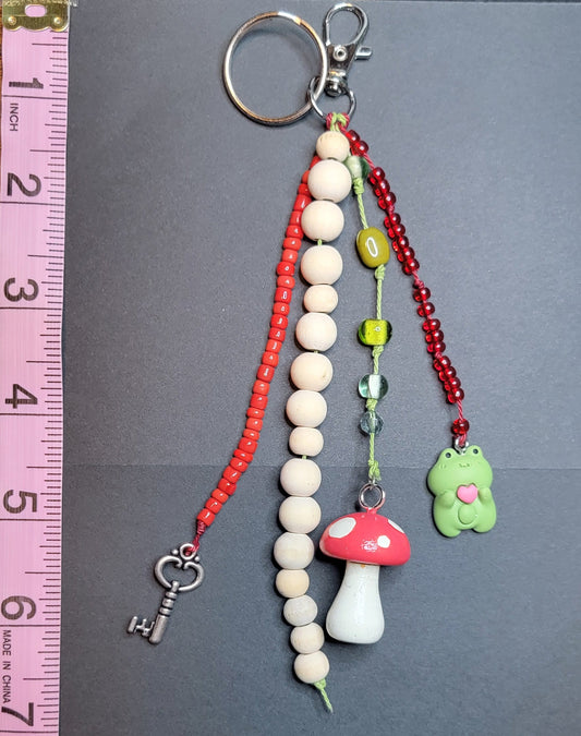 A handmade beaded keychain with charms, displayed on a dark gray card next to a pink measuring tape and a wooden table. The keychain features red and white beads, a white mushroom charm, a green flower charm, and a silver keyring.