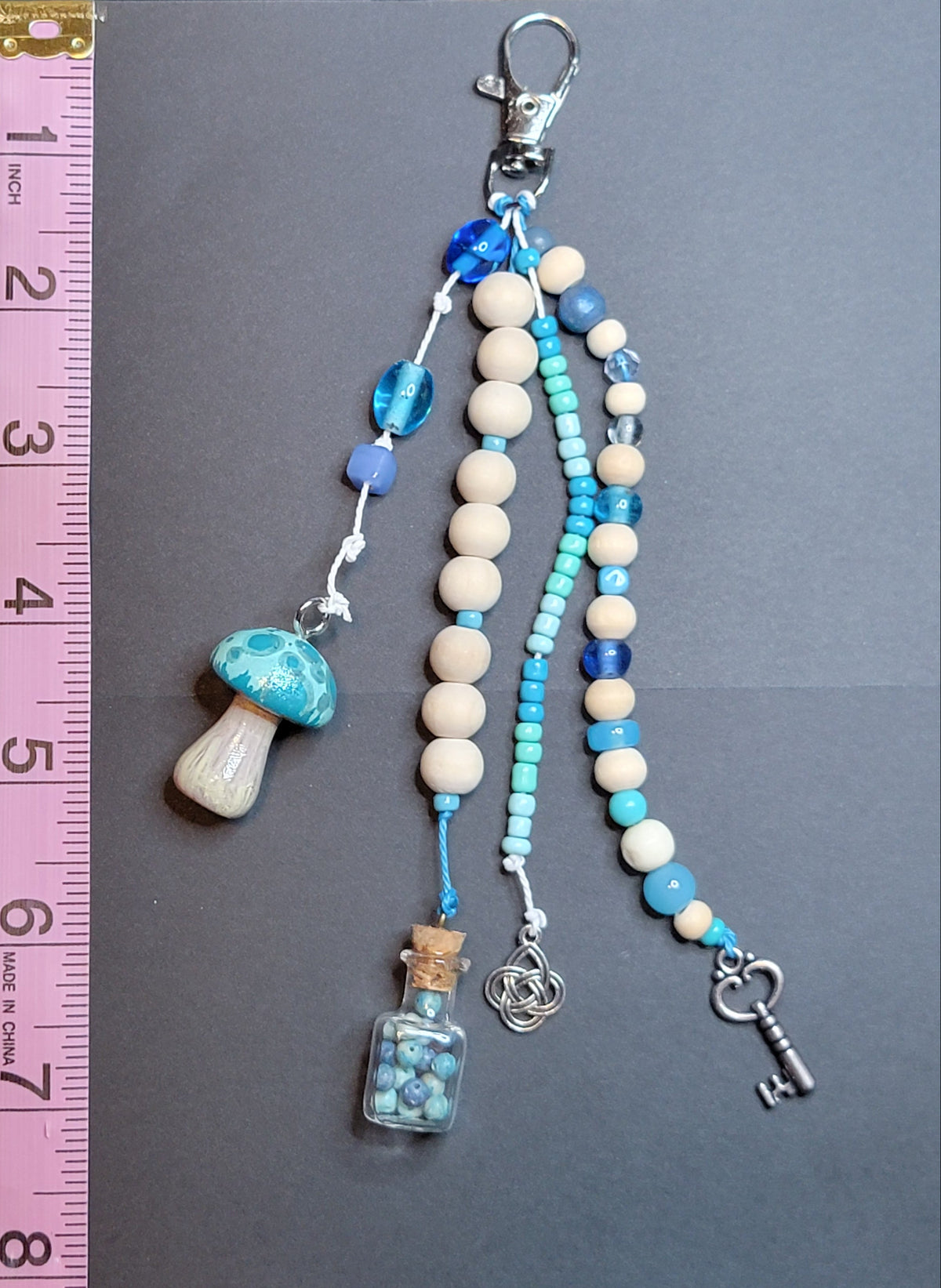 A handmade beaded keychain charm set displayed on a dark gray background next to a pink measuring ruler for scale. The keychain features cream-colored wooden beads, light blue and dark blue beads, a blue and white marbled mushroom charm, a small glass vial with blue beads, and a silver key.