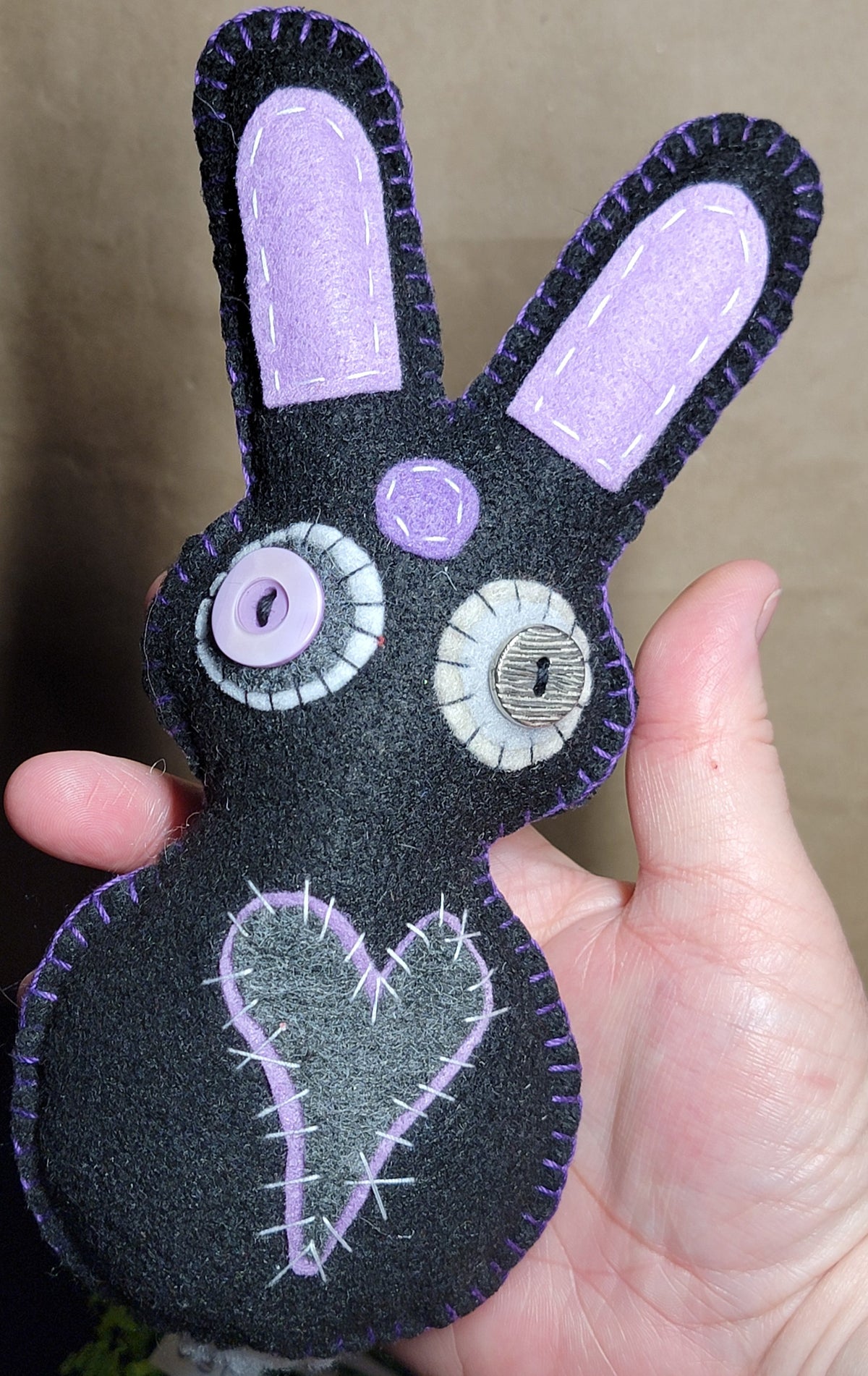 A handmade black felt bunny plush toy with lavender embroidered details, held in a person's hand against a neutral background.