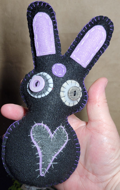 A handmade black felt bunny plush toy with lavender embroidered details, held in a person's hand against a neutral background.