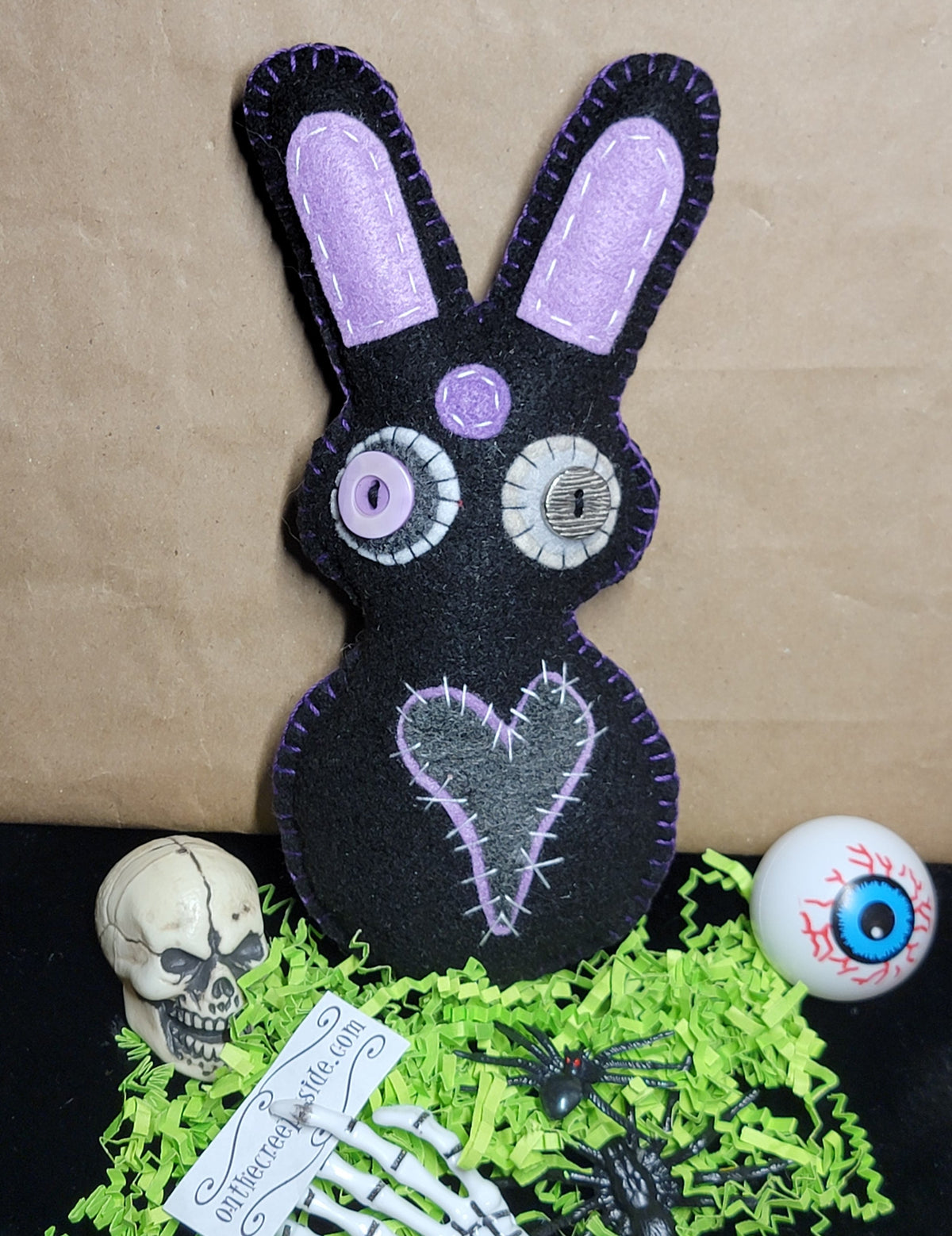 A black felt bunny decoration with lavender-purple ears, stitched eyes, and a stitched heart on the front, displayed on a black surface with green shredded paper, a small skull, a skeleton hand, black spiders, and a white eyeball prop against a brown paper background.