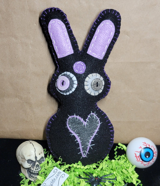 A black felt bunny decoration with lavender-purple ears, stitched eyes, and a stitched heart on the front, displayed on a black surface with green shredded paper, a small skull, a skeleton hand, black spiders, and a white eyeball prop against a brown paper background.