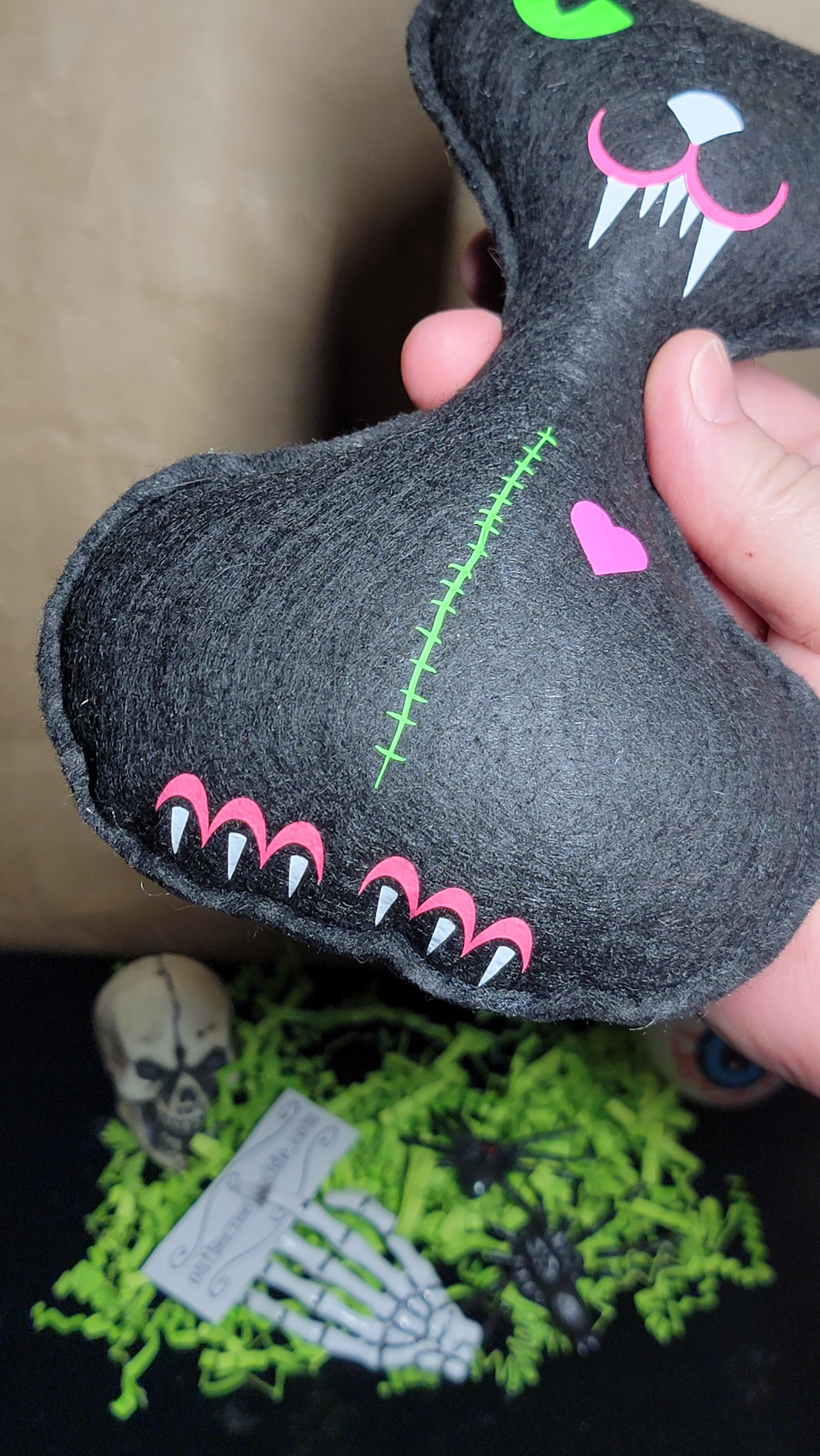 A hand holding a black felt monster-shaped object with pink, white, and green embroidered details, set against a dark background with Halloween props.