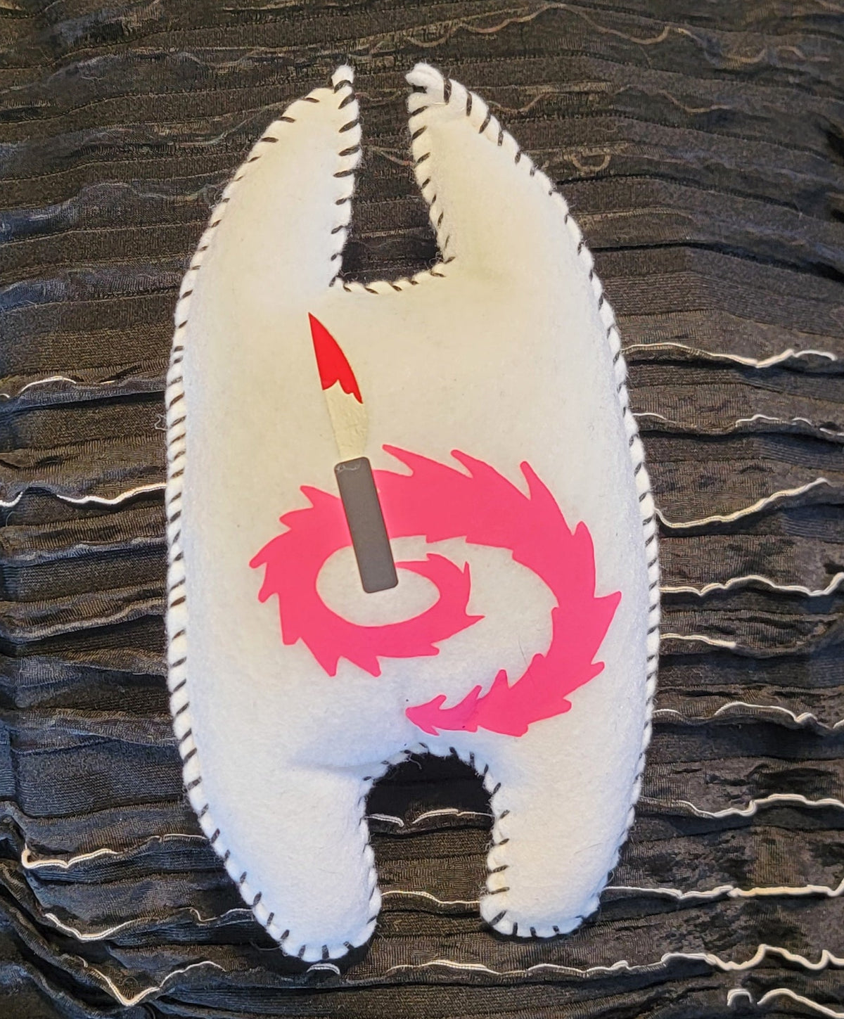 A black fringed fabric pouch or organizer with a white shell-shaped patch featuring a pink spiral and a red-and-black arrow, photographed indoors on a dark surface.
