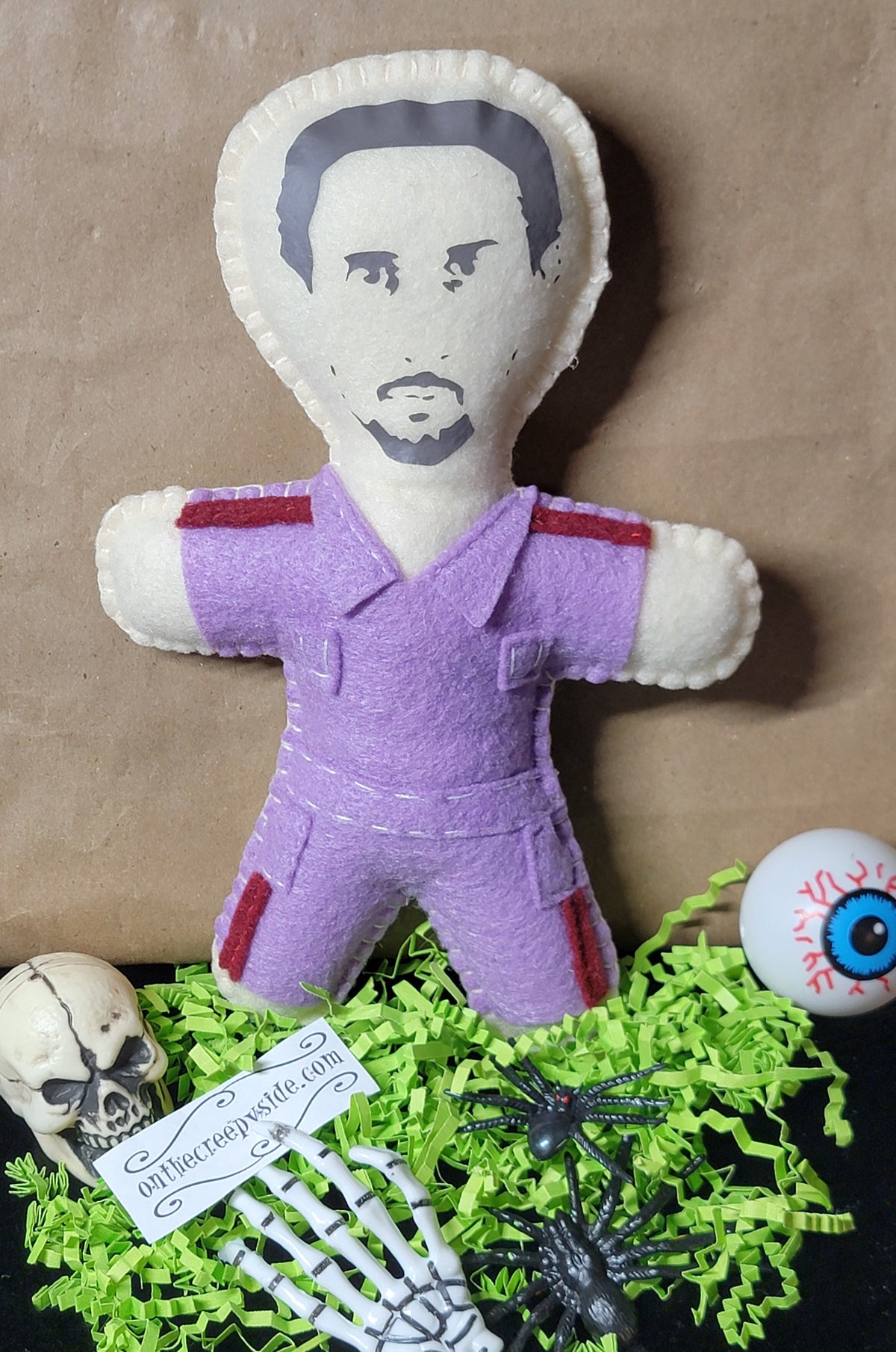 A handmade fabric doll with a printed face, wearing a purple jumpsuit with red accents, displayed on a black surface with green shredded paper, a skeleton hand, a skull, a spider, and a eyeball prop.