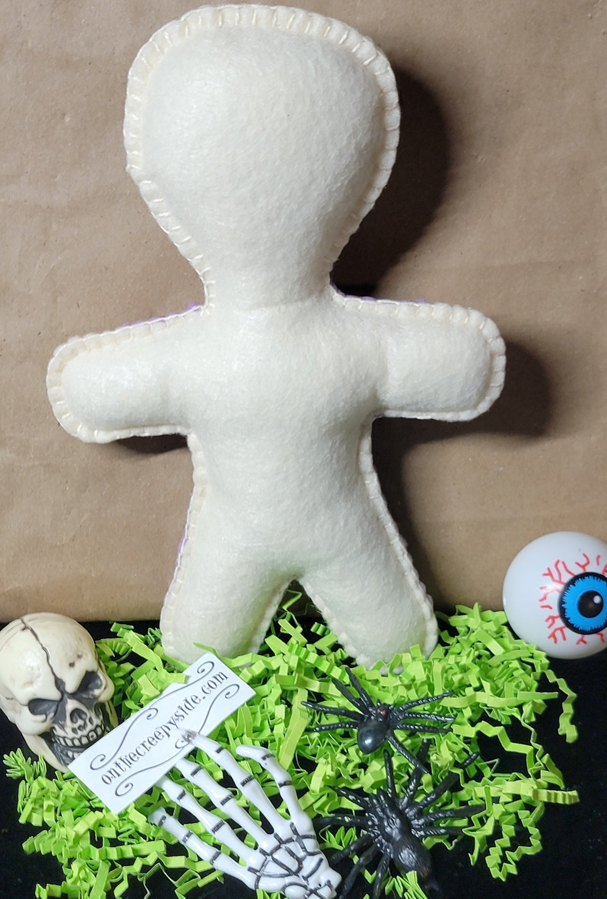 A white felt voodoo doll toy standing upright on a black surface, surrounded by Halloween props including green shredded paper, a small skull, a skeleton hand holding a tag, and a blue eyeball decoration.