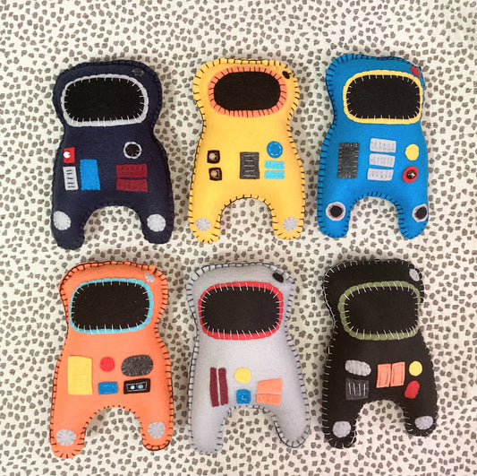 Six astronaut-shaped felt toys in different colors (navy, yellow, blue, orange, white, and black) arranged in two rows on a textured beige and gray patterned fabric background.