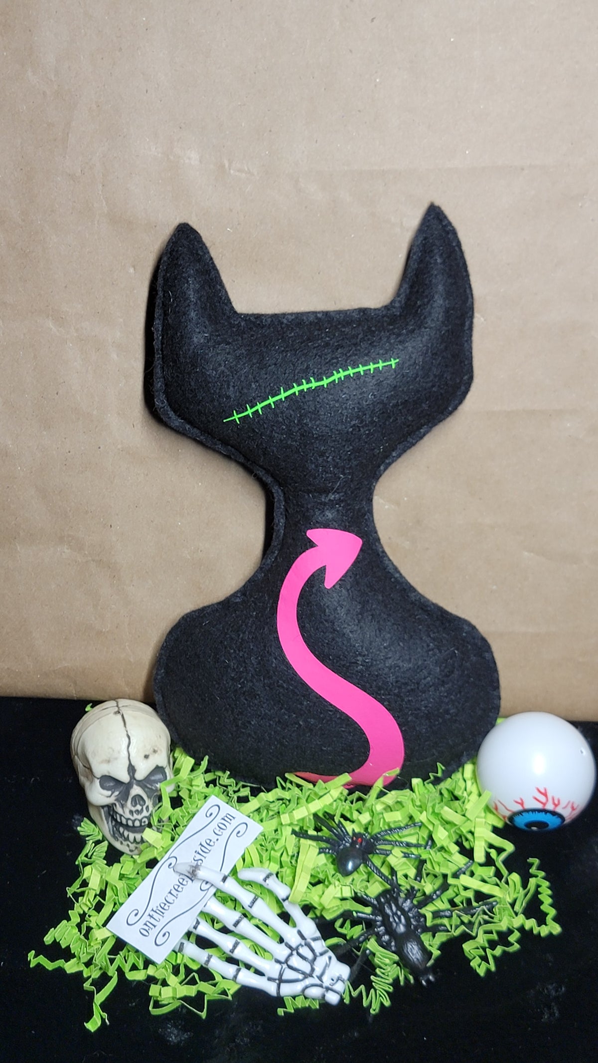 A black cat-shaped plush cushion with a pink arrow design and green stitched line details, displayed on a black surface with Halloween-themed props including a skull, skeleton hand, spiders, and a white eye ball against a brown paper background.