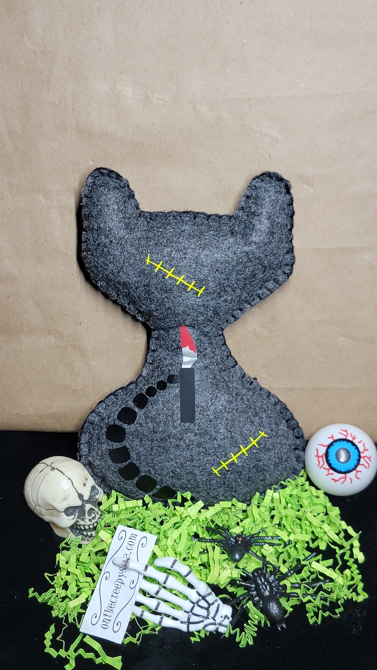 A Halloween-themed felt cat pillow in dark gray, styled on a black surface with green shredded paper, a small skull, a skeleton hand, a spider, a eyeball prop, and a ticket. The background is a brown kraft paper sheet.