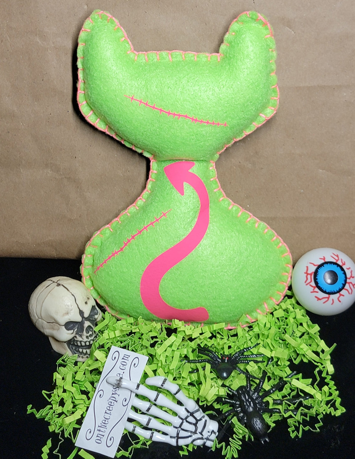 A bright green cat-shaped plush pillow with a pink arrow design on the front, displayed on a black surface with decorative Halloween props including a skull, skeleton hand, spider, eyeballs, and shredded green paper against a brown paper backdrop.