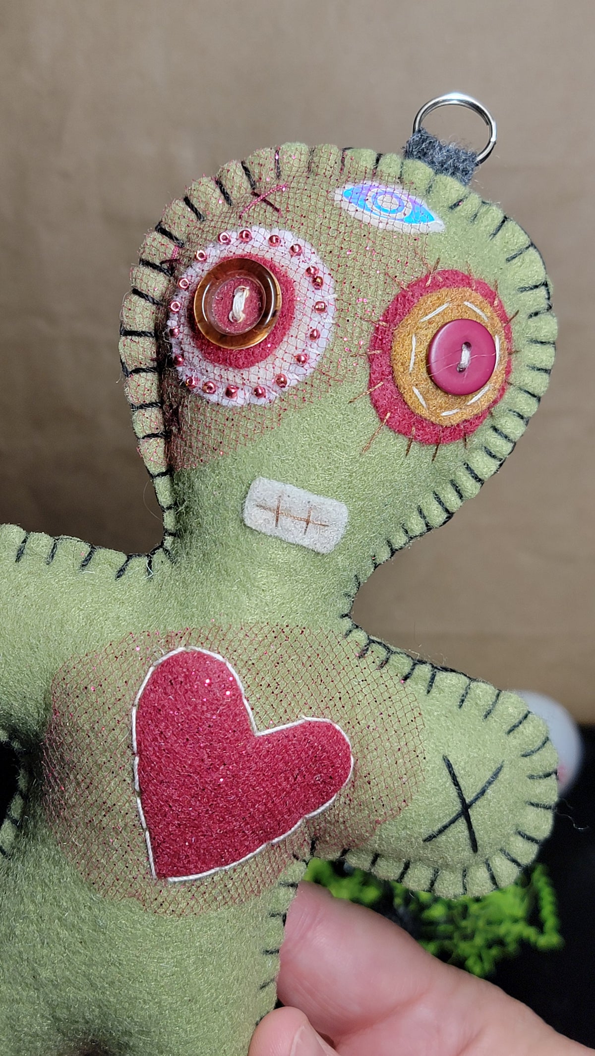 A handmade green fabric voodoo doll being held by a hand, featuring button eyes, a red glitter heart on the chest, and black stitched details.