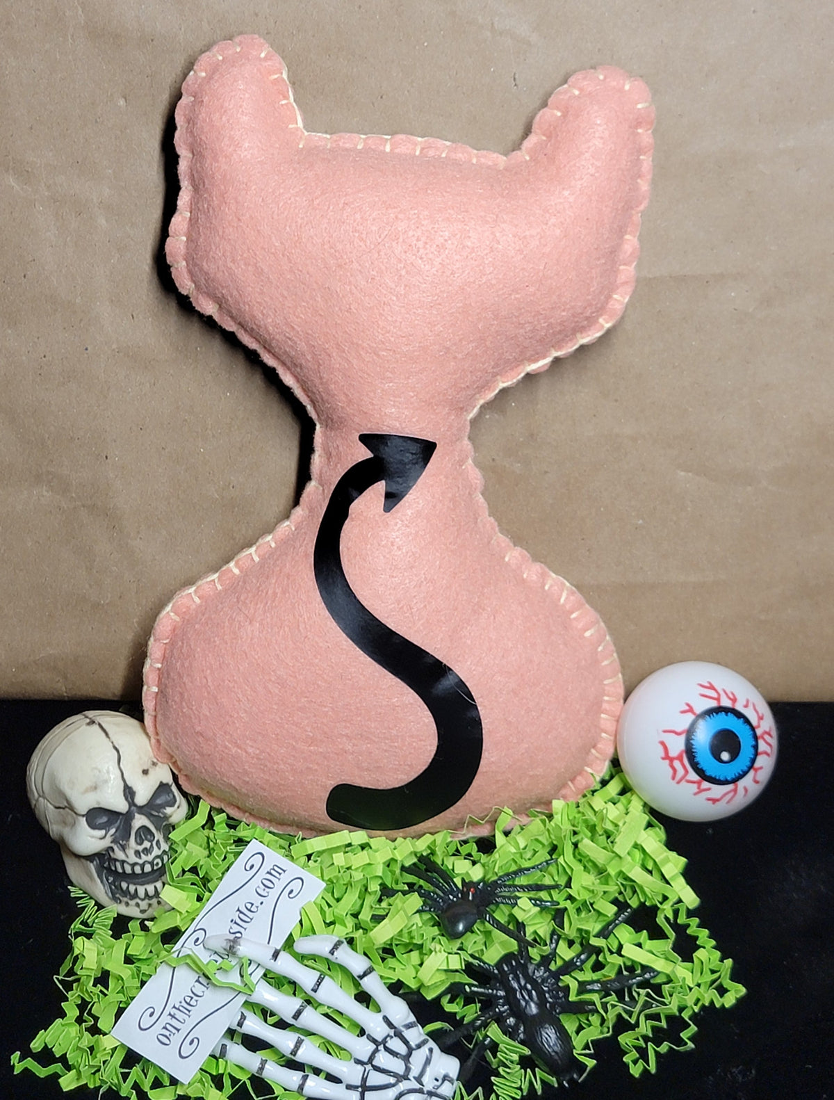 A pastel peac cat-shaped plush pillow with a black devil tail, displayed on a black surface with green paper shred, a skull, skeleton hand, spider, eyeball prop, and a brown kraft paper background.