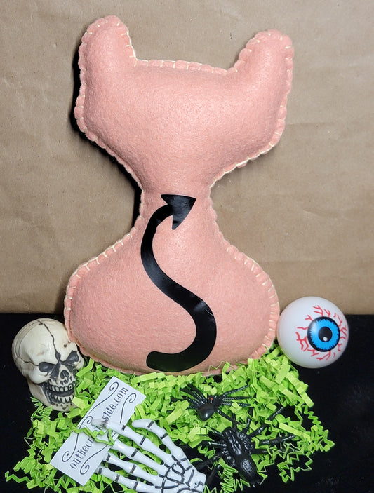 A pastel peac cat-shaped plush pillow with a black devil tail, displayed on a black surface with green paper shred, a skull, skeleton hand, spider, eyeball prop, and a brown kraft paper background.