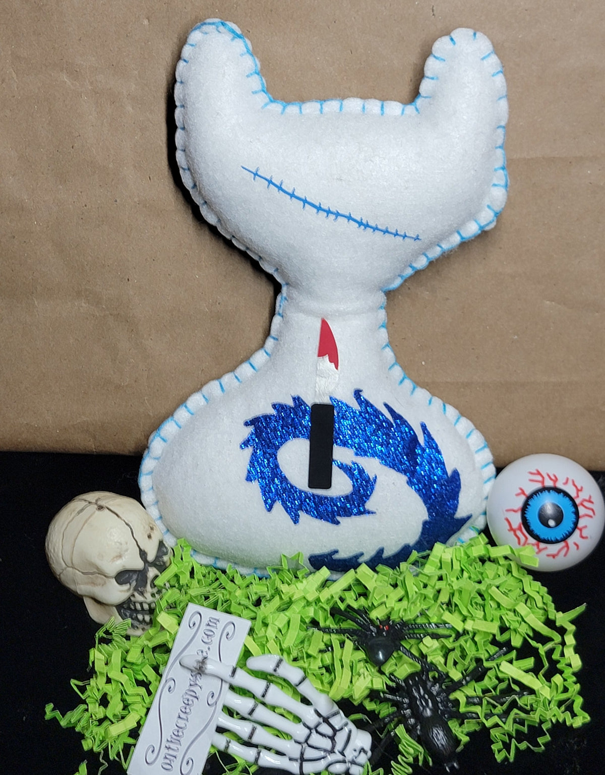 A white felt cat-shaped candle holder or decorative object with blue stitched trim, blue glittery swirl details, and a black candle inserted into the neck. It is displayed on bright green shredded paper with Halloween props including a small skull, a skeleton hand, a spider, and an eyeball against a brown paper background.