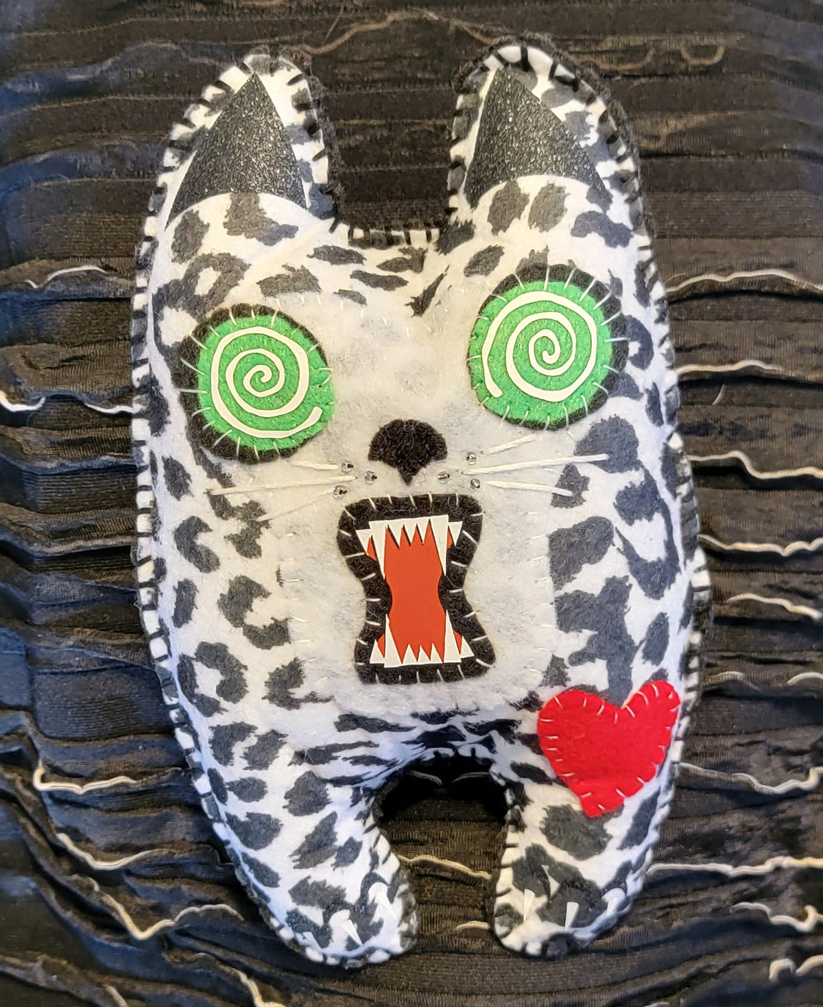 A handmade plush toy shaped like a cat head with a black and white spotted pattern, green spiral eyes, an open mouth showing red teeth, and a red heart on its cheek, resting on a black textured fabric background.