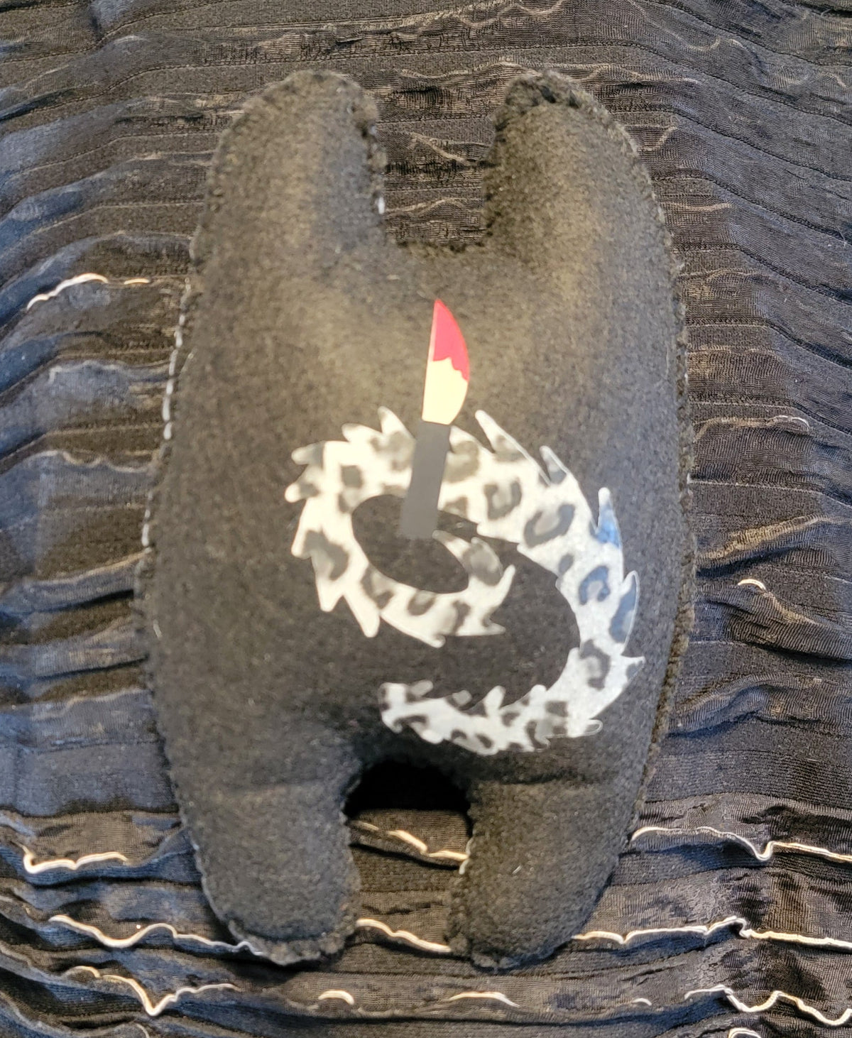 A black felt novelty pillow shaped like a rear end, featuring a white hand-drawn circle and a small red and white flag on the back, resting on a black and white wavy-patterned fabric background.