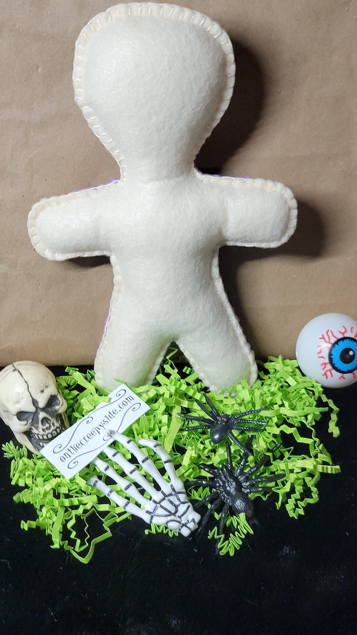 A white felt voodoo doll toy standing upright on a black surface, surrounded by Halloween props including green shredded paper, a small skull, a skeleton hand holding a tag, and a blue eyeball decoration.