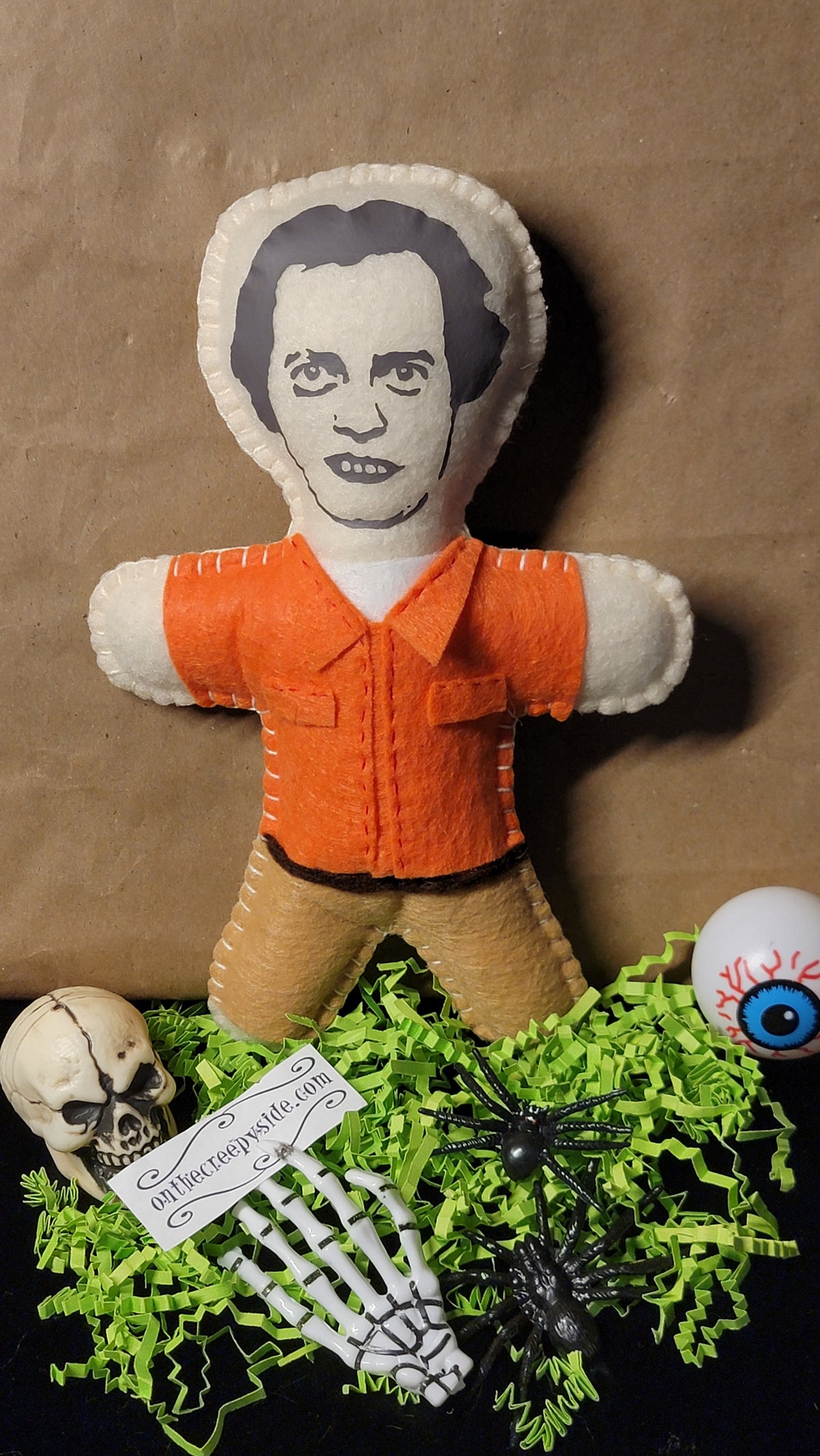 A handmade felt voodoo doll with a printed face, wearing an orange shirt and tan pants, styled on a brown background with skull, eye, skeleton hand, and black spider decorations.
