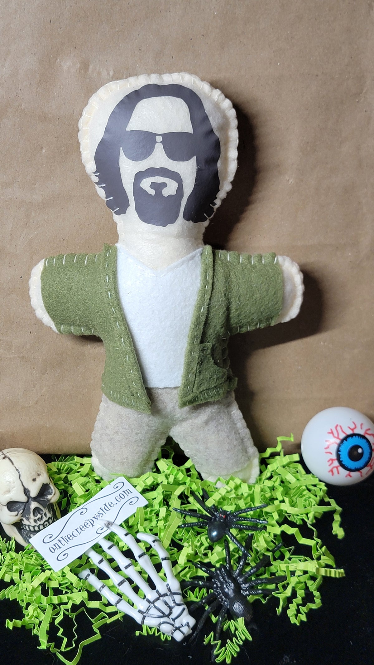 A handmade felt doll of a man with a beard and sunglasses wearing an olive green cardigan over a white shirt, styled with Halloween props including a skull, plastic skeleton hand, spiders, and a fake eyeball on a brown background.