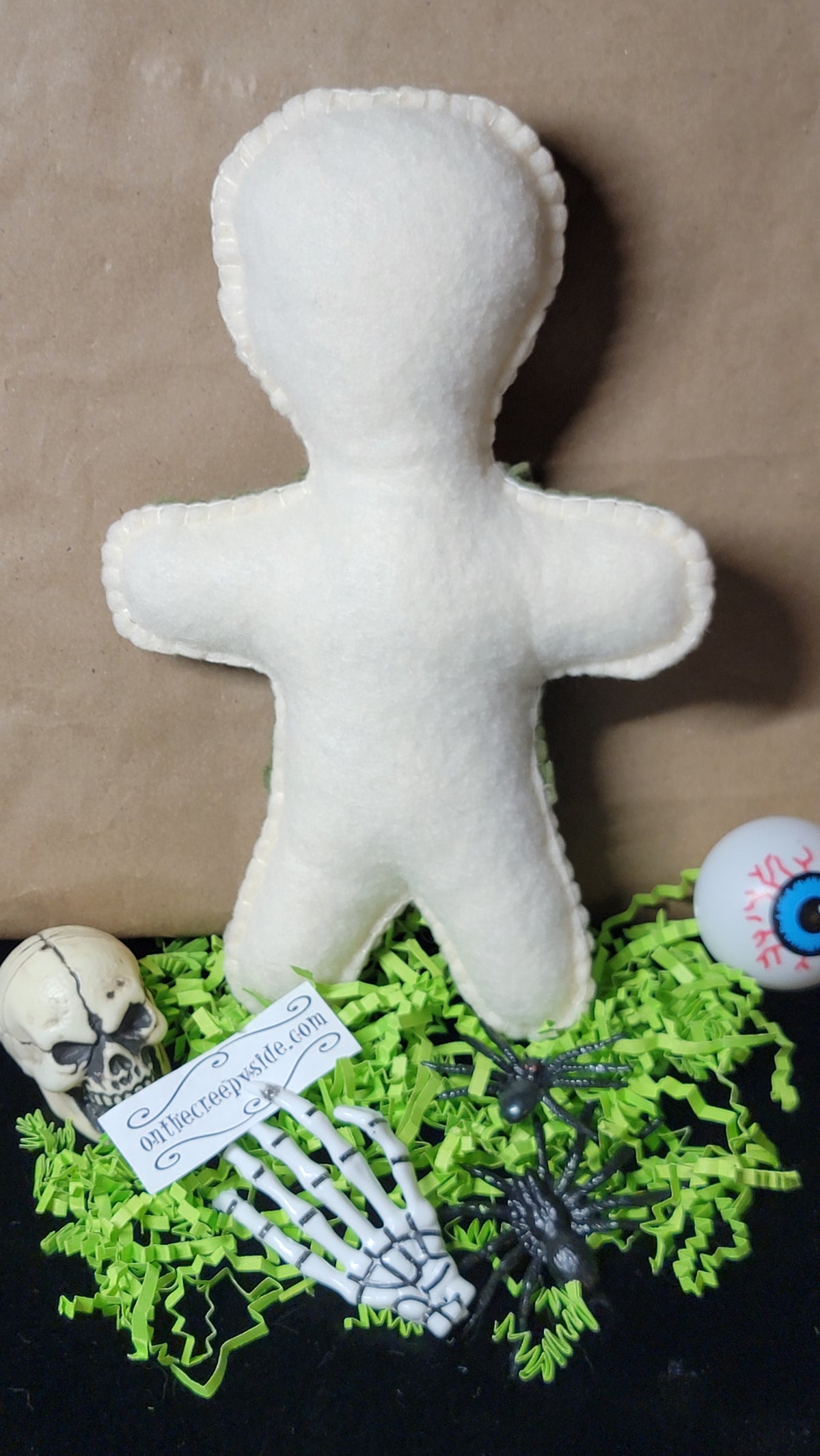 A white felt voodoo doll toy displayed on a black surface with green shredded paper, a small skull, skeleton hand, black spiders, and a white eyeball prop against a brown backdrop.