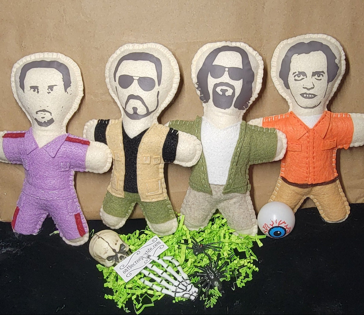 Four small fabric doll figures with printed faces and colorful outfits, arranged in front of a large brown paper bag on a black furry surface.