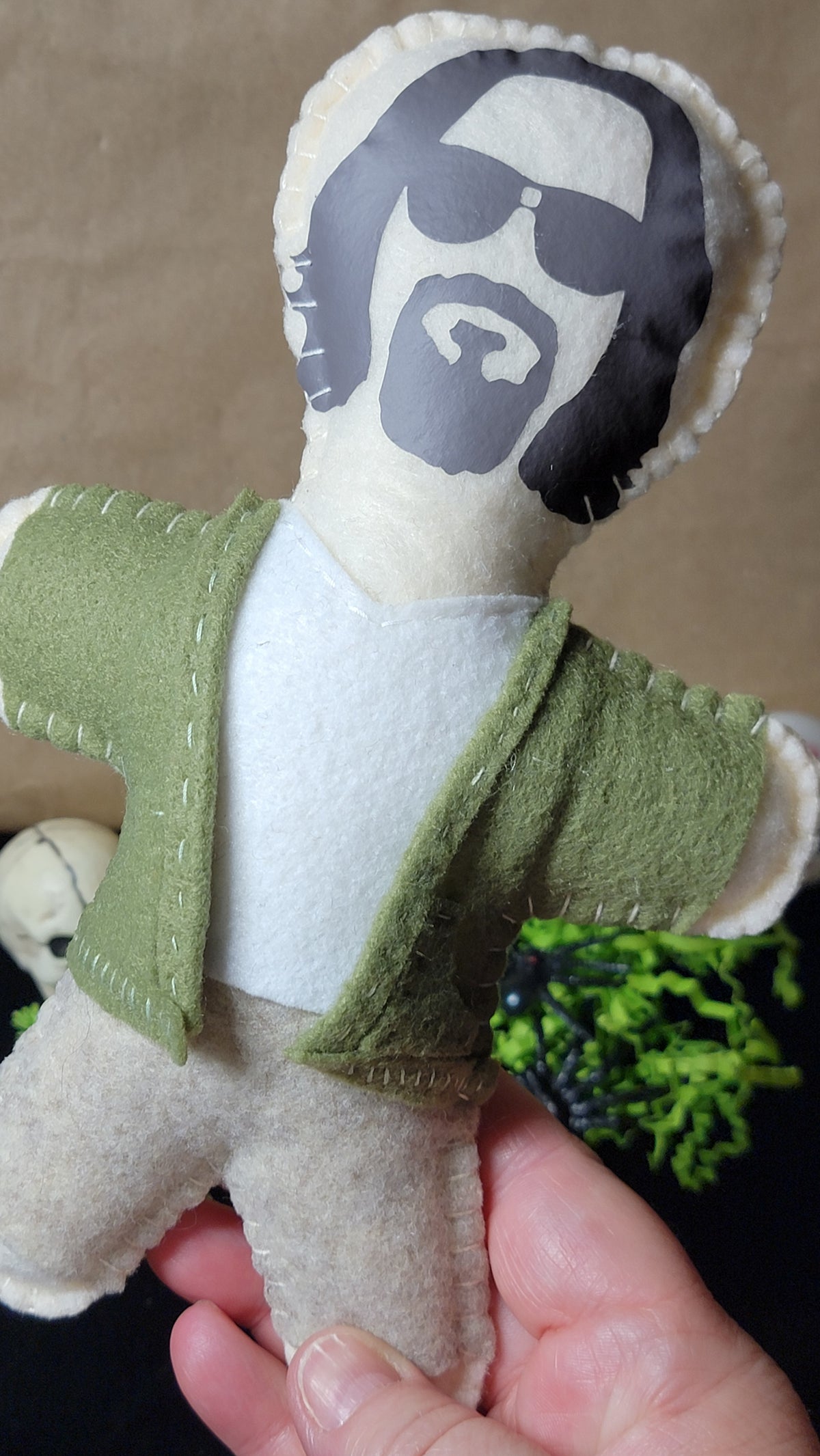 A handmade felt doll of Jesus Christ with a printed face, wearing an olive green robe over a white shirt, held in a person's hand against a brown background.