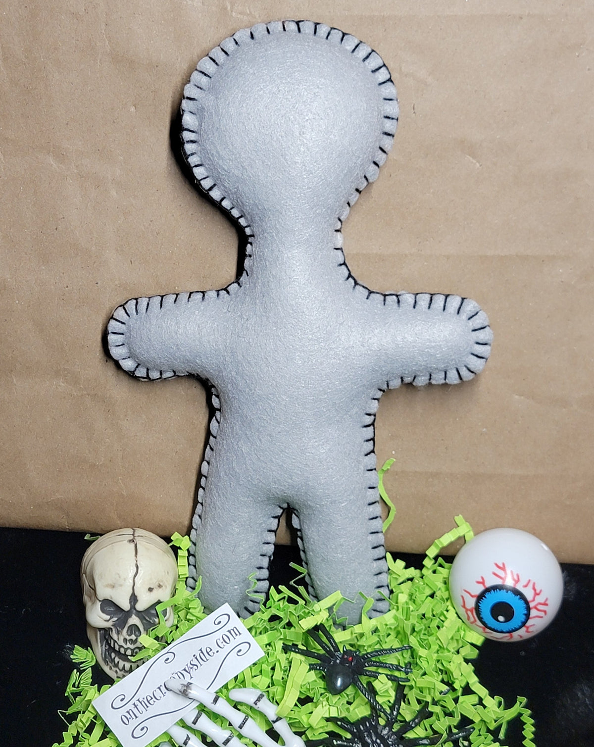 A gray fabric voodoo doll with black stitched edges, displayed on a black surface with green shredded paper, a small skull, a skeleton hand, a black spider, and a white eyeball prop against a brown paper background.