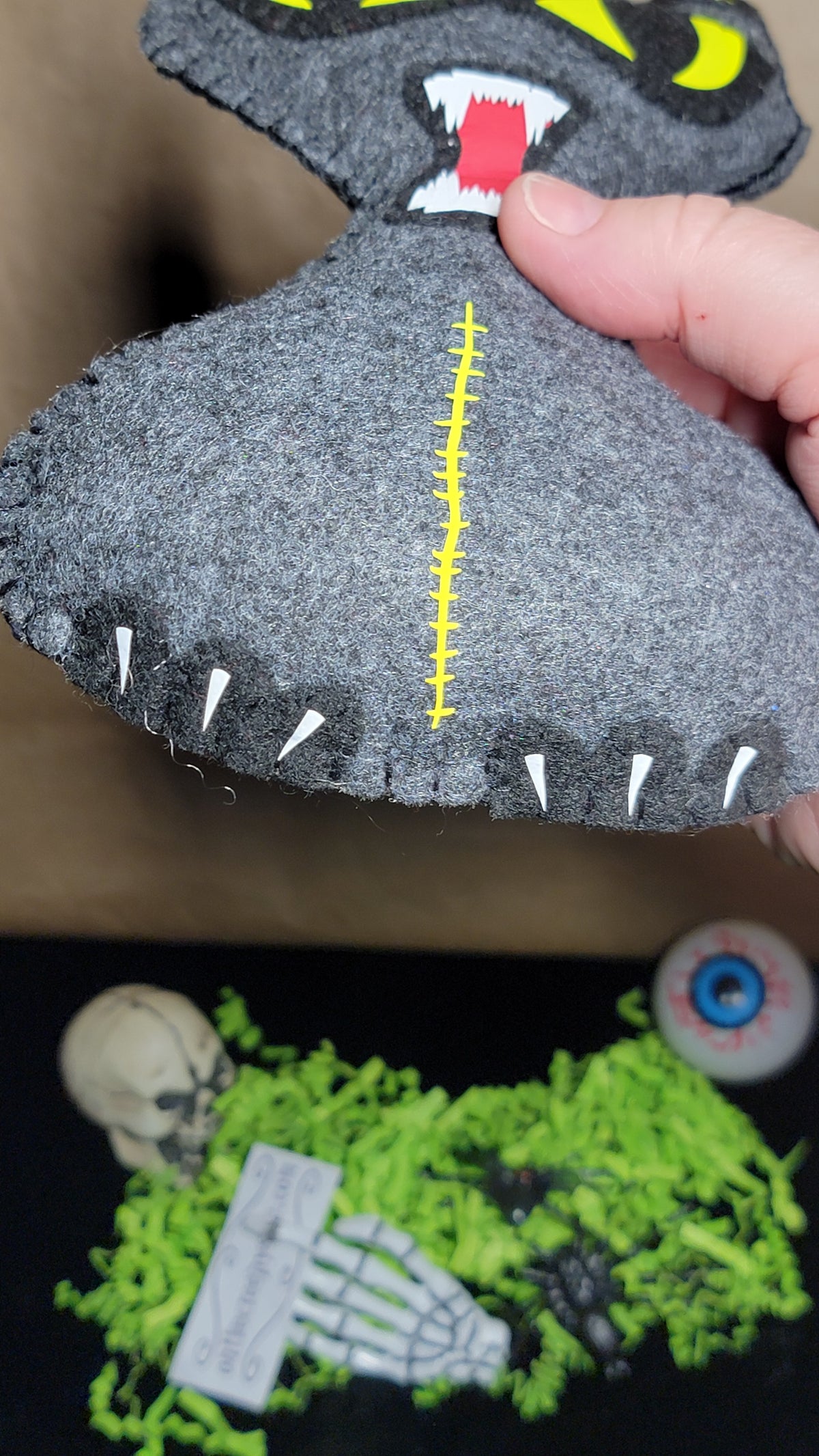 A hand holding a gray felt monster toy with yellow stitched details, white teeth, and a red tongue, shown against a dark background with other Halloween-themed decorations below.