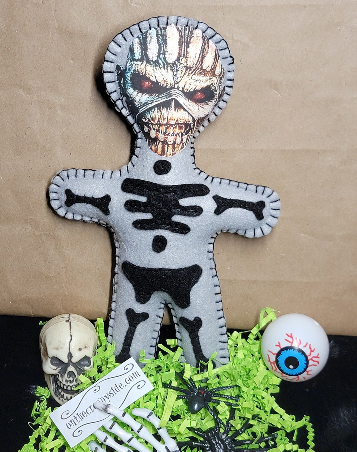 A skeleton-themed voodoo doll or plush figure made from gray felt, standing on green shredded paper with a skull, skeleton hand, black spiders, and a white eyeball prop against a brown paper backdrop.
