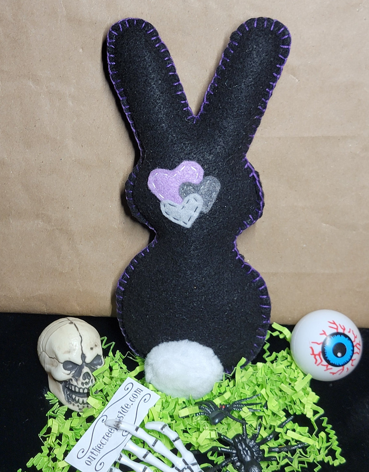 A black felt bunny plush toy with purple contrast stitching, featuring a pink and gray heart design on its chest. The bunny is displayed on a black surface with decorative Halloween props, including a small skull, a white eyeball, a skeleton hand, a spider, and green shredded paper, against a brown kraft paper background.