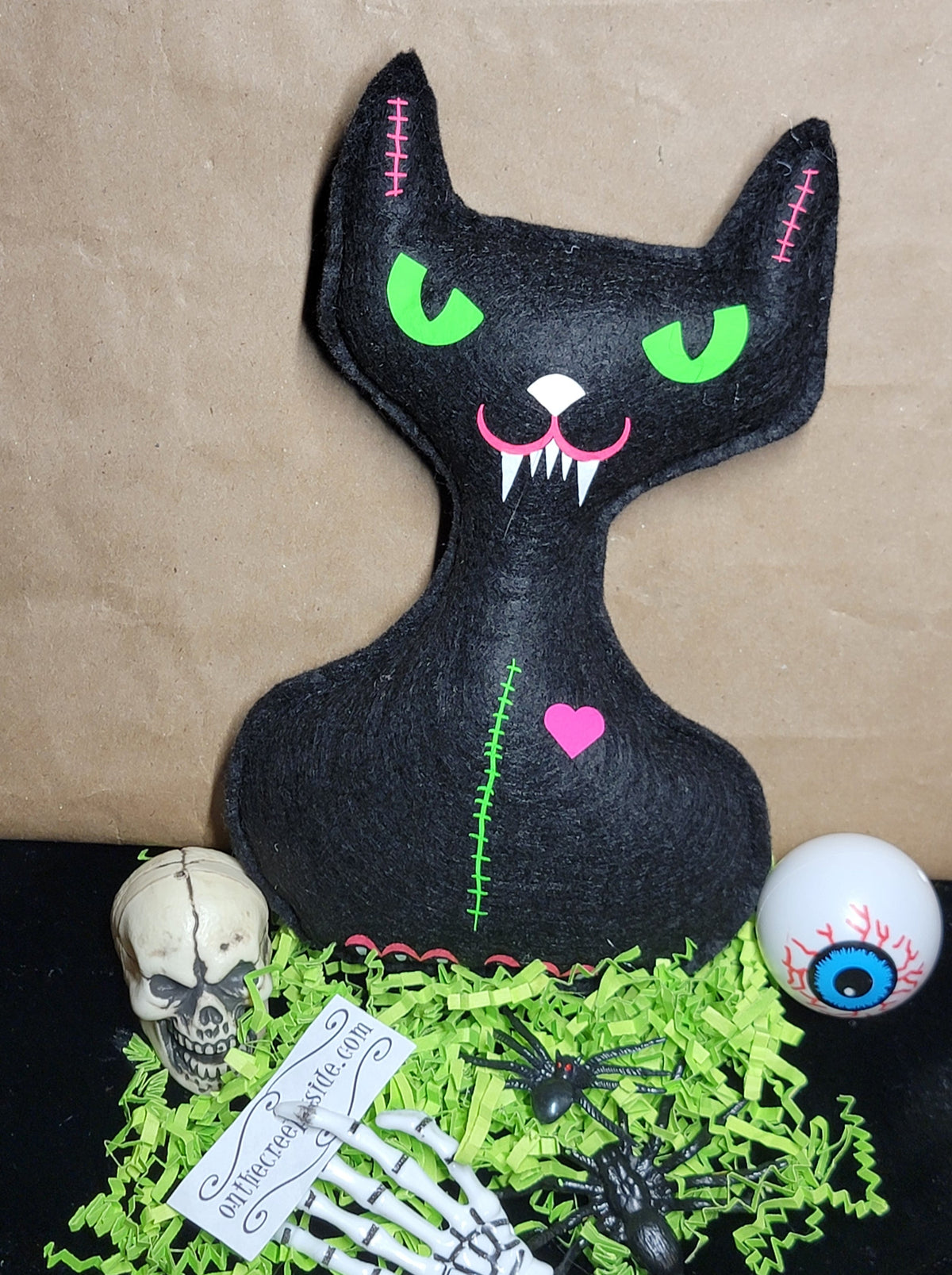 A black cat-shaped Halloween decoration with bright green eyes, pink stitched details, and a pink heart on the chest, displayed on a black surface with green paper shred, a skull, skeleton hand, spiders, and an eyeball prop against a brown paper backdrop.