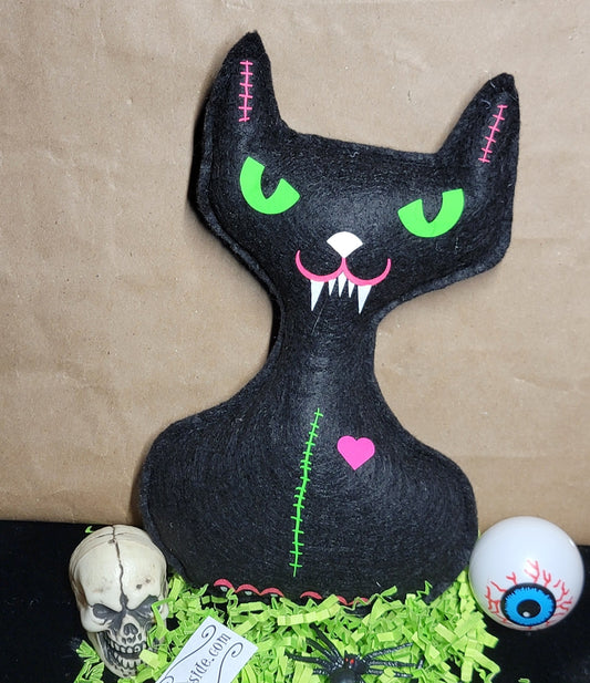 A black cat-shaped Halloween decoration with bright green eyes, pink stitched details, and a pink heart on the chest, displayed on a black surface with green paper shred, a skull, skeleton hand, spiders, and an eyeball prop against a brown paper backdrop.