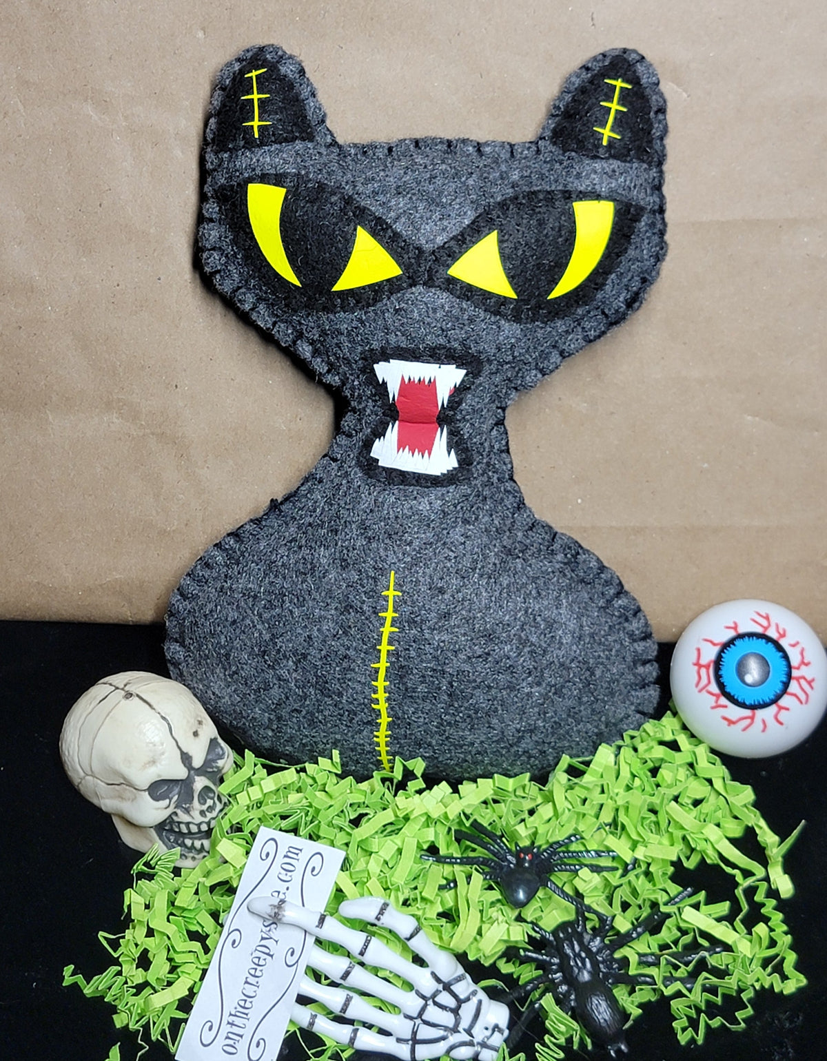 A black felt cat plush toy with glowing yellow eyes and an open mouth showing white teeth, styled on a black surface with green shredded paper, a small skull, a skeleton hand, a spider, a tag, and a gory eyeball prop against a brown paper background.