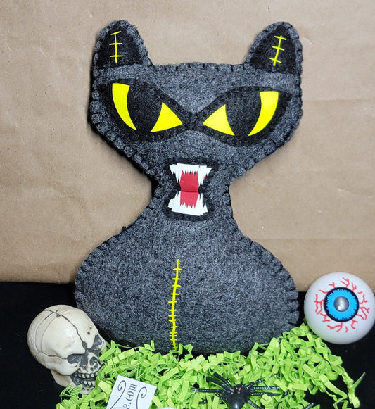 A black felt cat plush toy with glowing yellow eyes and an open mouth showing white teeth, styled on a black surface with green shredded paper, a small skull, a skeleton hand, a spider, a tag, and a gory eyeball prop against a brown paper background.