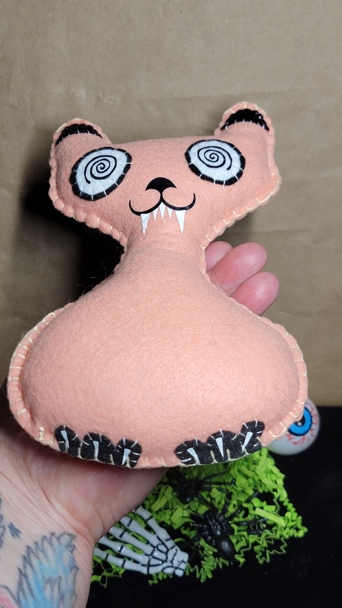 A handmade peach-colored felt cat plush toy being held by a hand. The cat has black and white spiral eyes, a stitched black nose and mouth, white fangs, and black-and-white striped paws.