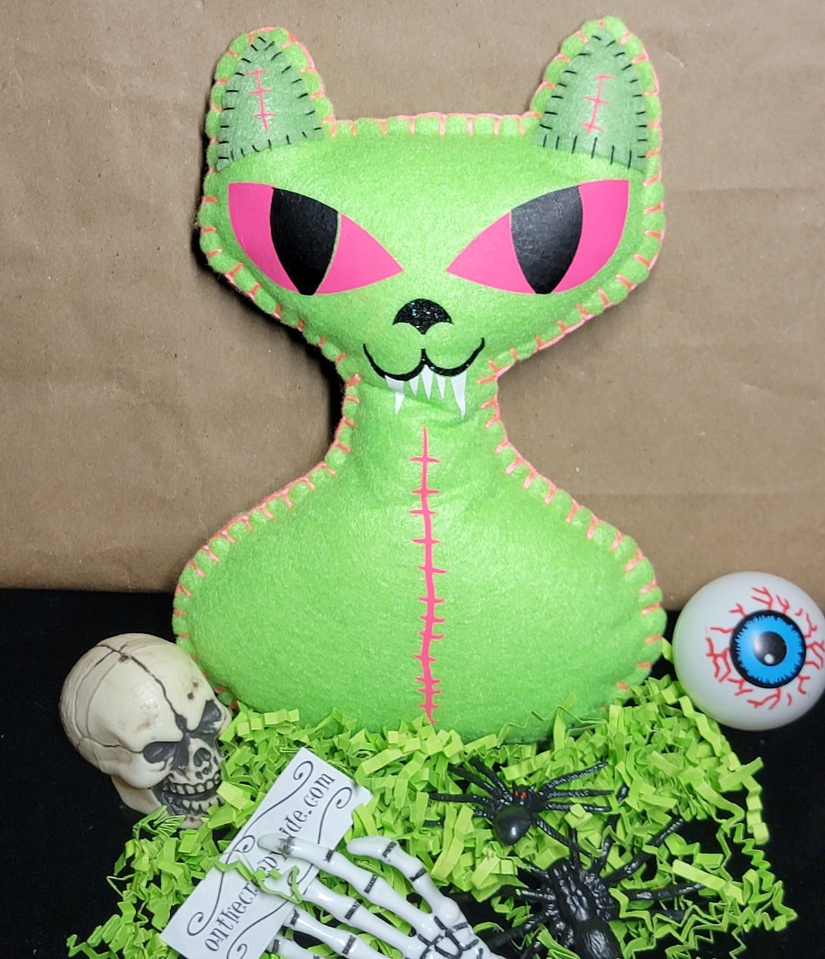 A green felt cat-shaped Halloween decoration with pink and black embroidered eyes and mouth, displayed on a black surface with shredded green paper, a small skull, a skeleton hand, a spider, and an eyeball prop against a brown paper backdrop.