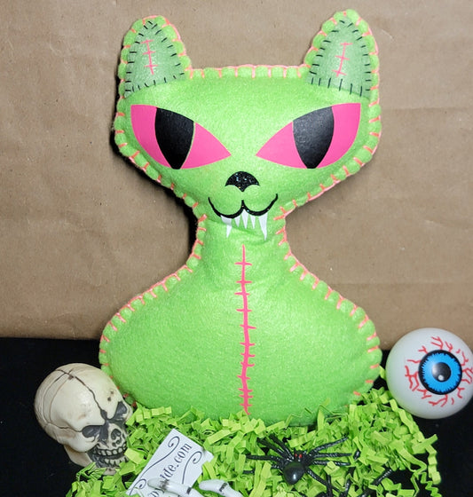 A green felt cat-shaped Halloween decoration with pink and black embroidered eyes and mouth, displayed on a black surface with shredded green paper, a small skull, a skeleton hand, a spider, and an eyeball prop against a brown paper backdrop.