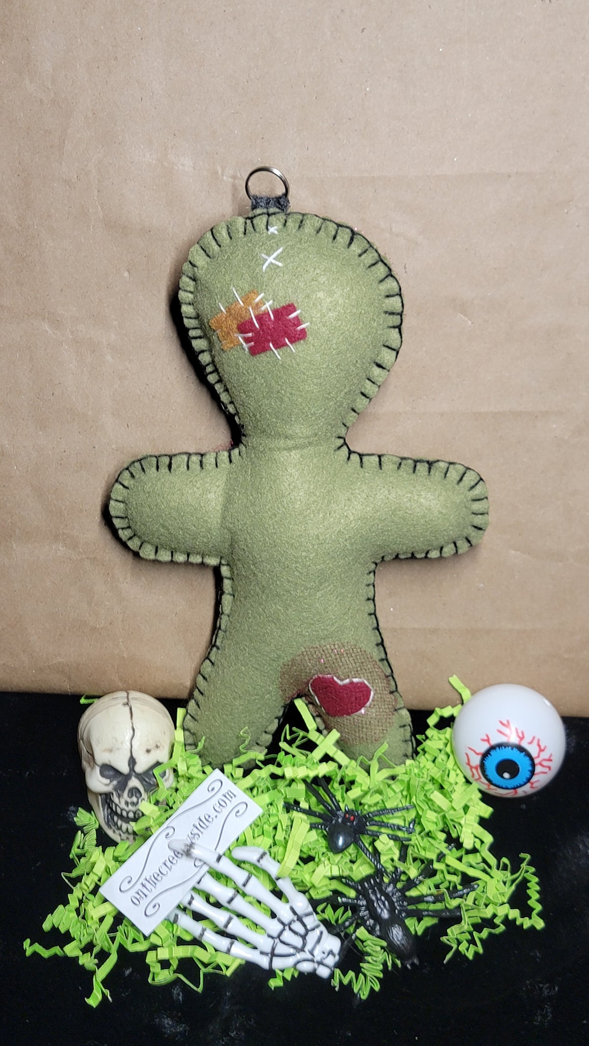 A handmade olive green felt voodoo doll hanging against a brown kraft paper backdrop, styled with a small skull, white eye ball, skeleton hand, black spiders, and bright green shredded paper on a black surface.