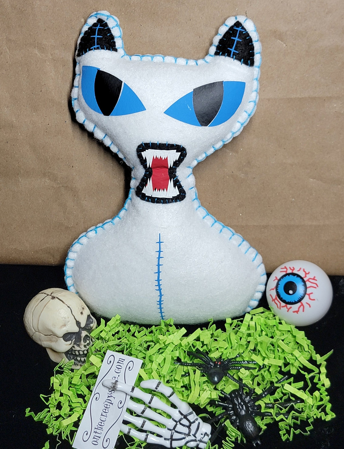 A white plush cat pillow with blue eyes and stitched details, displayed on a black surface with green shredded paper, a small skull, a眼球 ornament, and skeleton accessories against a brown paper backdrop.