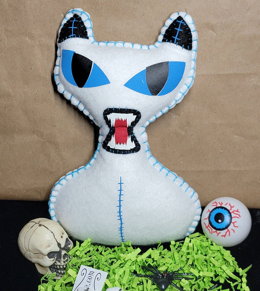 A white plush cat pillow with blue eyes and stitched details, displayed on a black surface with green shredded paper, a small skull, a眼球 ornament, and skeleton accessories against a brown paper backdrop.