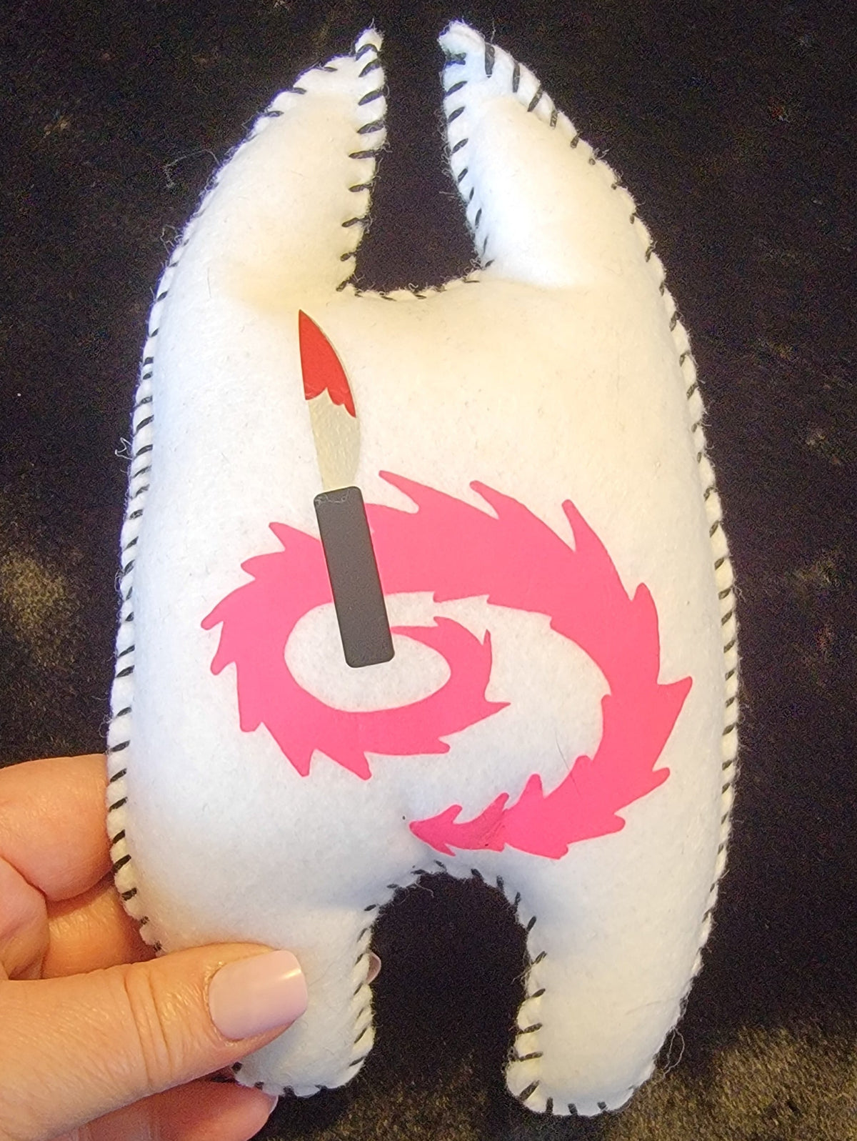 A hand holding a white felt tooth-shaped plush toy with black stitched edges, featuring a pink spiral design and a small red-and-black lipstick graphic on the front, set against a dark textured background.