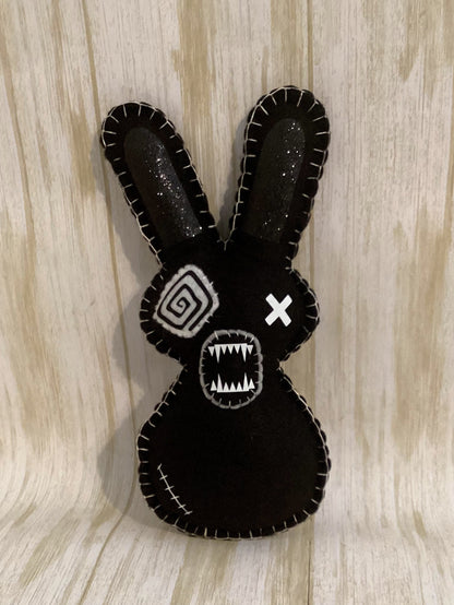 A black felt rabbit head decoration with white stitched details, featuring one spiral eye, one X eye, and a toothy grin, set against a light wood grain background.