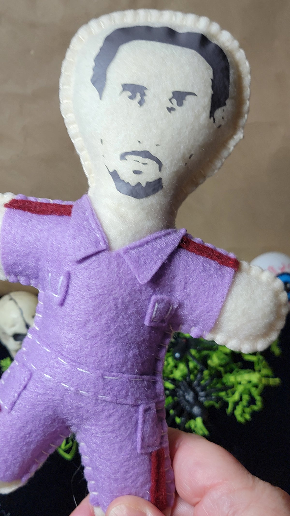 A handmade felt doll with a printed face, wearing a purple jumpsuit with red trim, held in a hand against a neutral background.