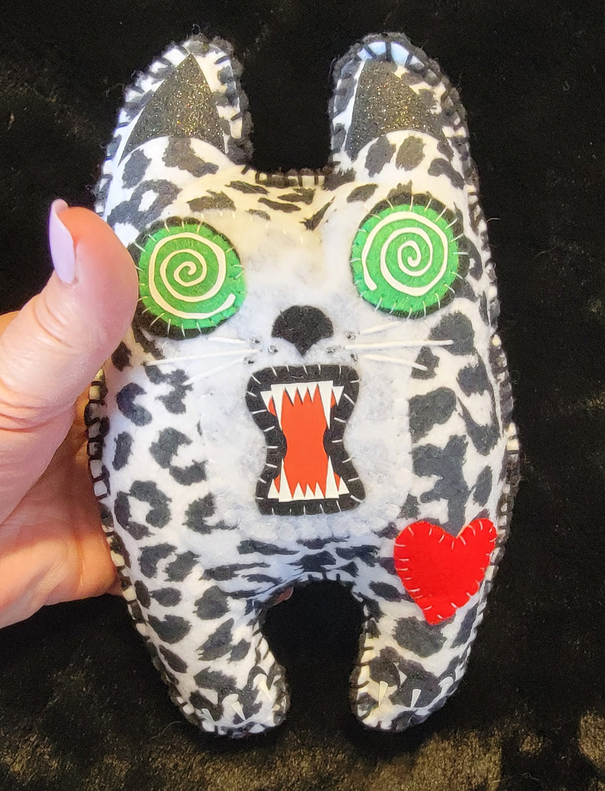 A hand holding a plush felt animal toy with a black and white spotted pattern, green spiral eyes, an open mouth showing red and white teeth, and a red heart on a dark textured background.