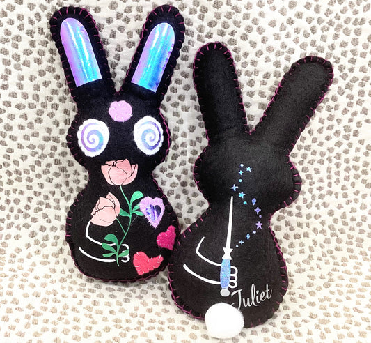 Front and back views of a black Magic Love Bunny plush with spiral eyes, holographic ears, flowers, hearts, and a personalized wand design.
